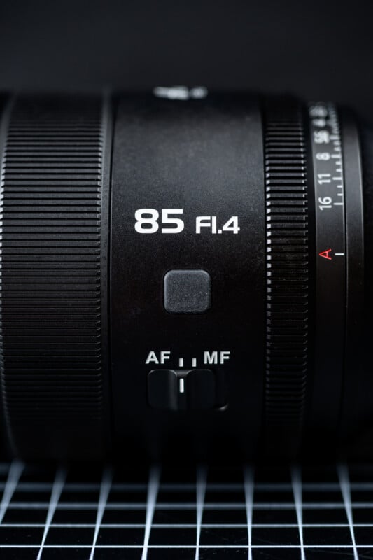 Close-up of a black camera lens with "85 F1.4" marked in white text, showing the AF/MF switch and part of the focus ring, placed on a surface with a white grid pattern.