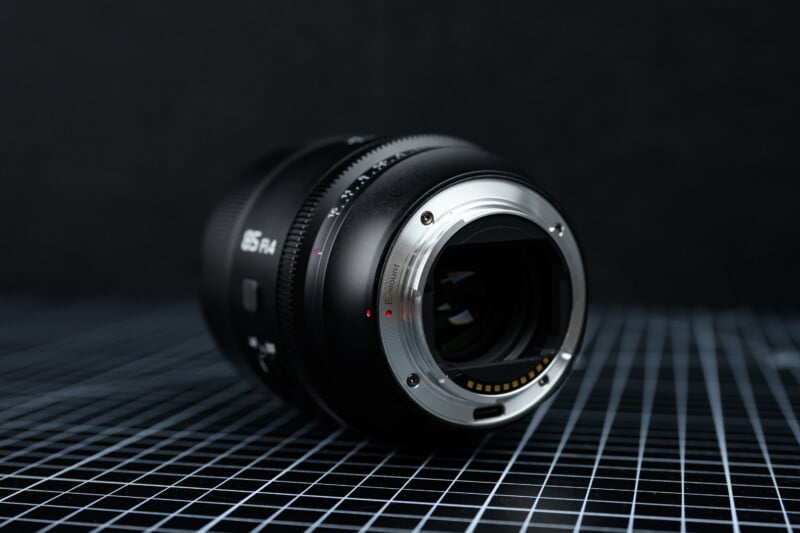 A camera lens positioned on its side atop a black grid-patterned surface, with the metal mounting ring and electronic contacts facing the viewer. The background is dark and out of focus.
