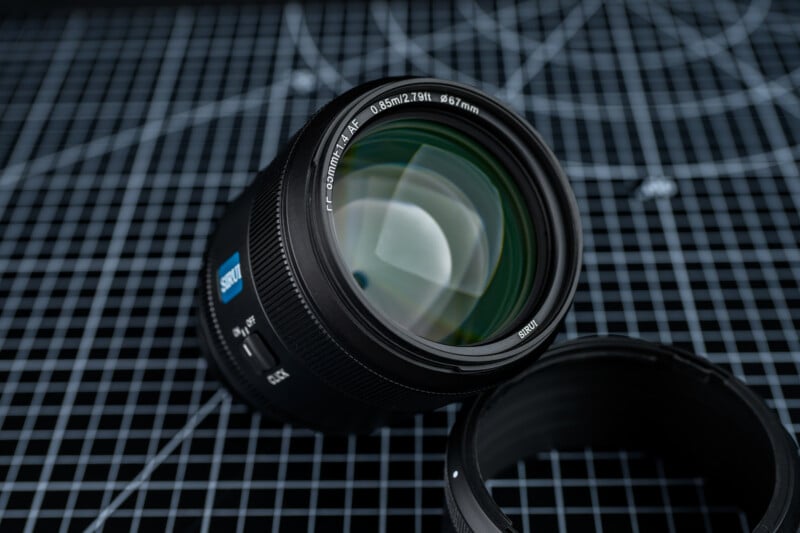 A close-up of a camera lens with "ZEISS" branding placed on a black grid-patterned surface, next to a lens hood. Lines and reflections are visible on the glass surface.