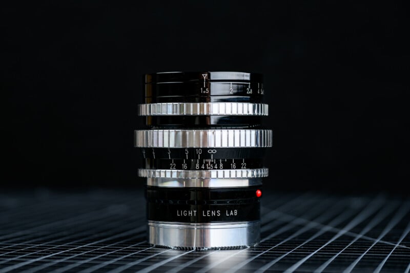 A close-up of a shiny, black and silver camera lens labeled "Light Lens Lab," standing upright on a grid-patterned surface with a dark, blurred background.