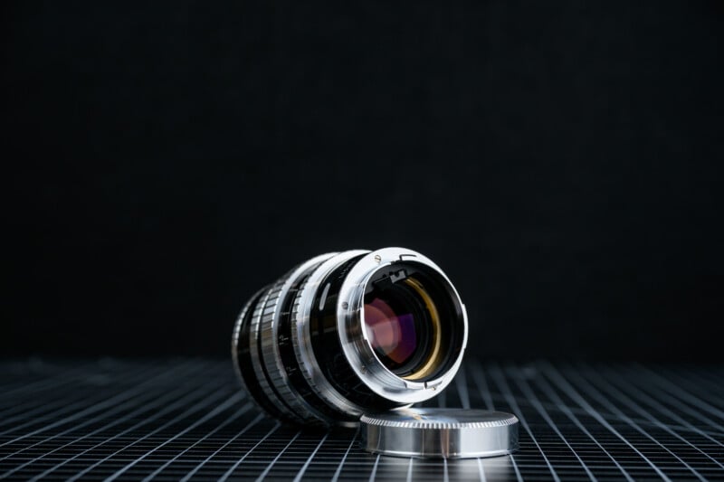 A camera lens lies on its side on a black grid-patterned surface, with its metallic lens cap placed nearby. The background is dark and out of focus, highlighting the lens as the main subject.