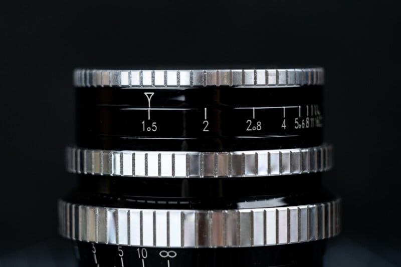 Close-up of a camera lens showing silver focus and aperture rings, with numbers and markings in white on a black background. The focus is on the aperture scale, featuring values from 1.5 to 8.