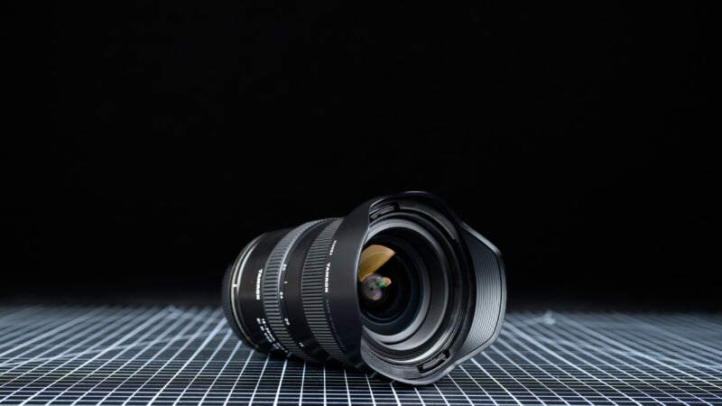 A wide-angle camera lens lies on its side atop a blue and white grid surface, with a dark black background.