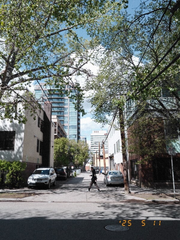 A person walks across a quiet urban street lined with parked cars, low-rise buildings, and tall trees. Modern high-rise buildings are visible in the background. The date "’25 5 11" is stamped in the corner.