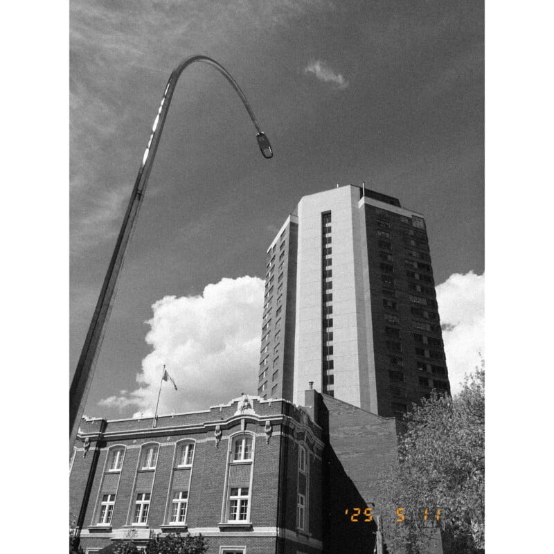 A black-and-white photo of a tall modern building behind an older brick structure, with a curved streetlamp in the foreground and clouds in the sky. A date stamp reads "’23 5 14" in the lower right corner.