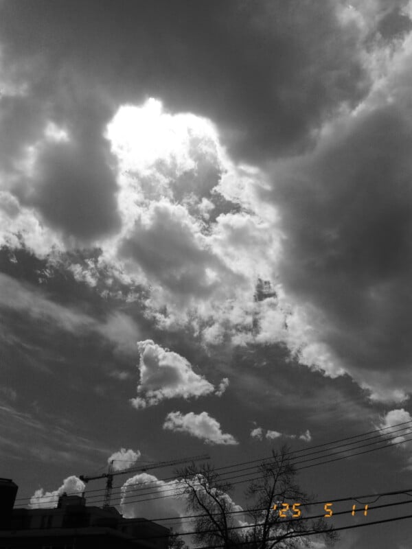 A dramatic sky filled with large, bright clouds partially obscuring the sun, above silhouettes of trees, power lines, and buildings. The date “25 5 11” appears in orange at the bottom right corner.