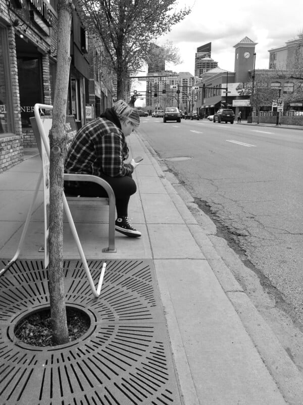 A person wearing a plaid jacket sits on a bus stop bench, looking at their phone. The street beside them is quiet, with parked cars and shops. A small tree grows beside the bench, and tall buildings are visible in the background.