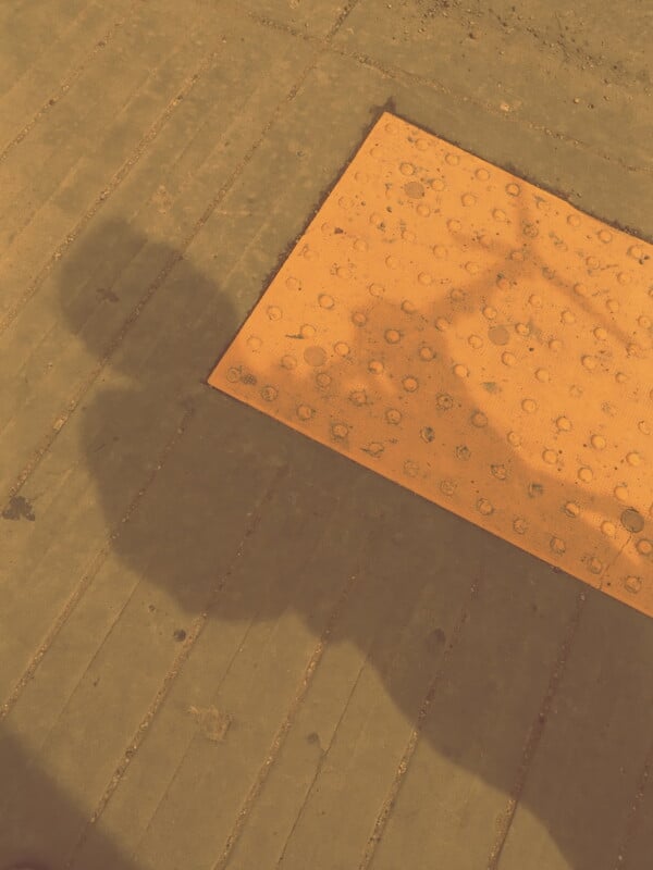 A shadow of a person holding something is cast on a sidewalk next to a yellow textured tactile paving tile with raised dots, typically used for visually impaired accessibility. The scene appears to be outdoors in sunlight.