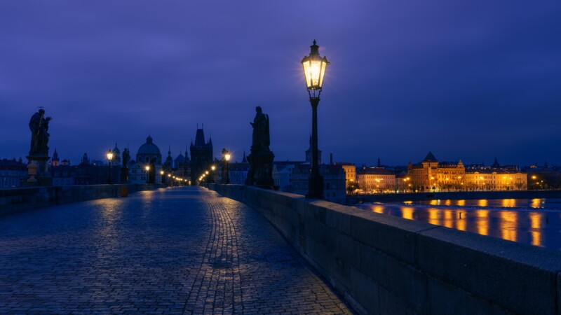 A cobblestone bridge under a dark blue sky, illuminated by street lamps. Statues line the path, with a distant city skyline featuring lit buildings reflecting on the water.