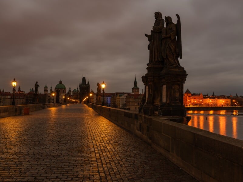 Stone statues stand on a dimly lit, cobblestone bridge at dusk. Warm streetlights illuminate the path, with historic buildings and a distant city skyline under a cloudy sky on the horizon.