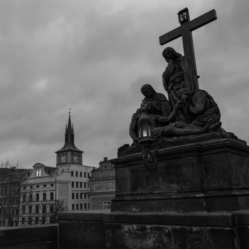 A black and white image of a religious statue with a cross, depicting several figures in a solemn pose. In the background, a historic building with a clock tower is visible under a cloudy sky.