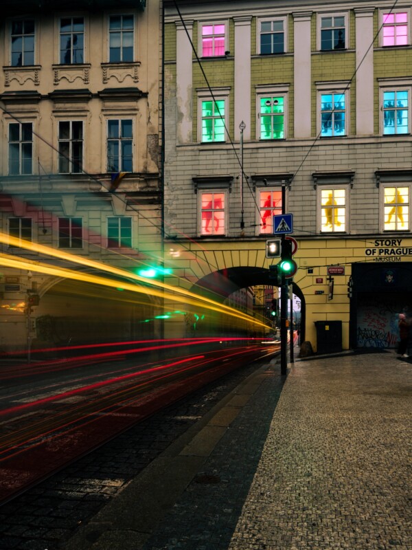 A street scene at night with a blurred passing tram leaving light trails. A building displays multicolored lights in its windows, casting a vibrant glow. A cobblestone sidewalk and crosswalk sign are visible in the foreground.