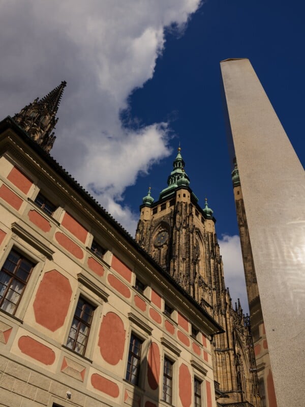 A dramatic view of a historic building with intricate architecture, featuring tall spires under a partly cloudy blue sky. In the foreground, a tall, modern obelisk reflects sunlight.