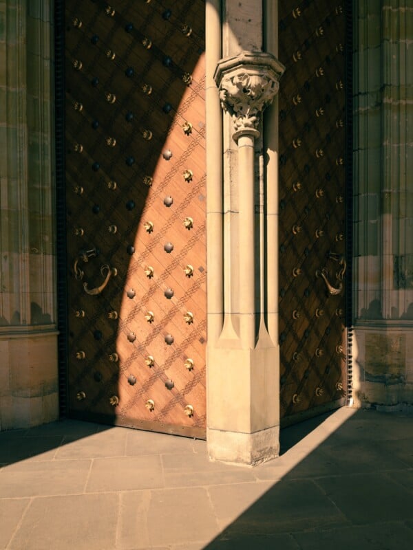 A large wooden door adorned with ornate metal studs and rings is partially open, casting a shadow on the stone floor. The door is framed by intricate stone architecture with a decorative column.