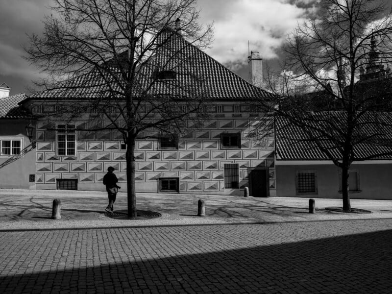 Black and white image of a solitary figure walking on a cobblestone path in front of a patterned building. Bare trees cast shadows on the ground. The scene has a calm, historic ambiance.