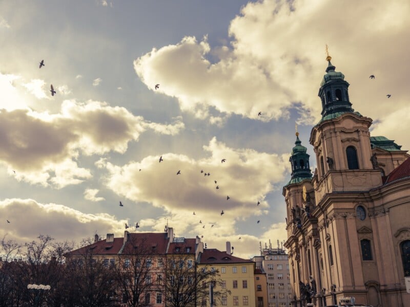 Historic European cityscape with Baroque architecture. A large church with green-domed towers stands next to pastel-colored buildings. Birds fly in a partly cloudy sky, and bare trees are visible, suggesting late autumn or early winter.