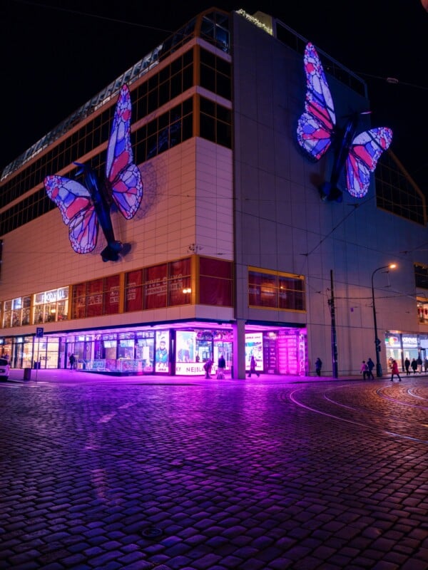 A building at night with large illuminated butterfly decorations on the exterior. The street and walkway glow with purple-pink light reflections. Bright storefront windows showcase various advertisements. Cobblestone street leads to a corner.