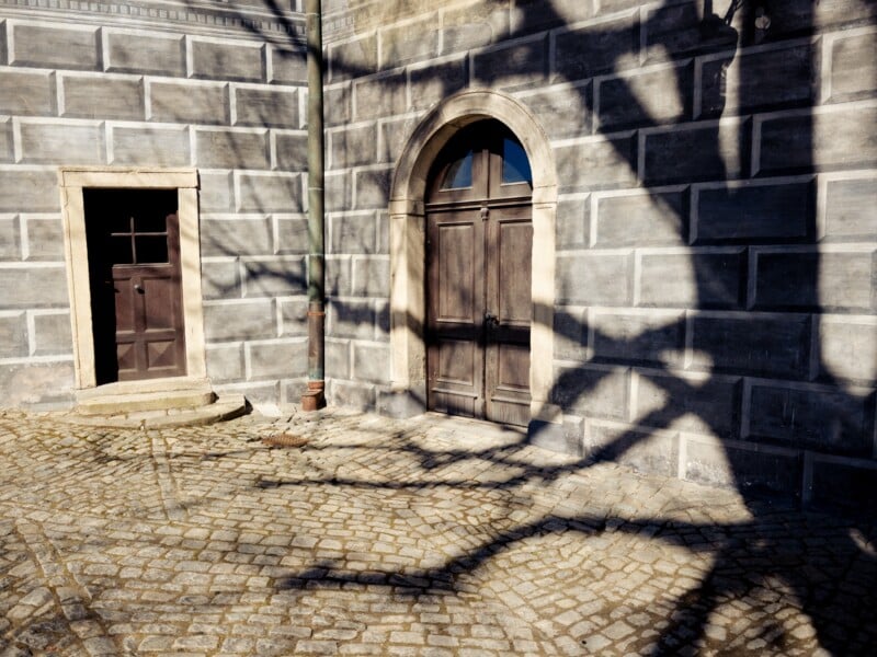Shadow of a tree on a stone building with two wooden doors. The ground is paved with cobblestones, creating a textured walkway in the sunlight. The walls feature a rectangular stone pattern.