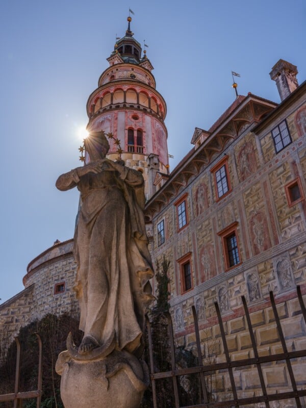 A stone statue stands in the foreground with an ornate historical tower and building behind it. The sun is partially visible, creating a halo effect around the tower's top. The building features intricate architectural details and decorative elements.