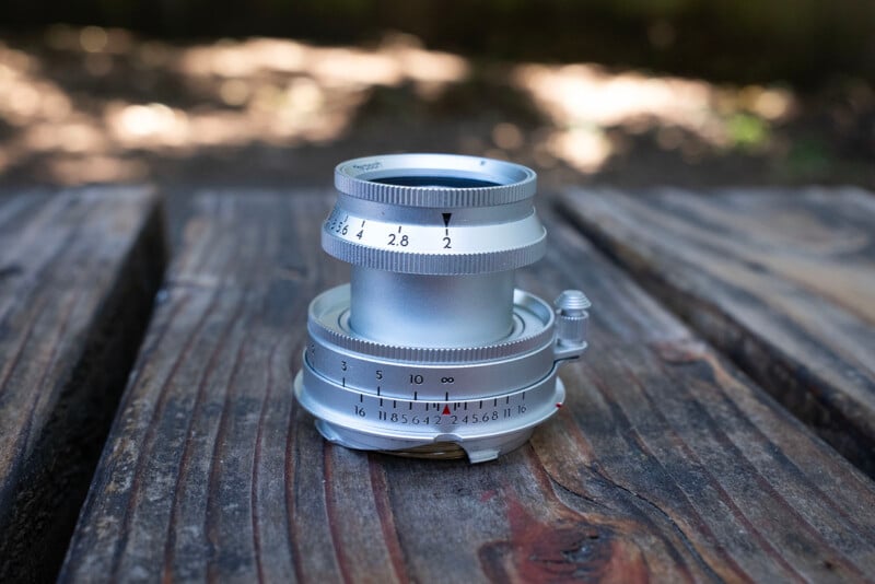 A silver camera lens with engraved numbers sits on a weathered wooden surface outdoors, with a blurred background of trees and sunlight.