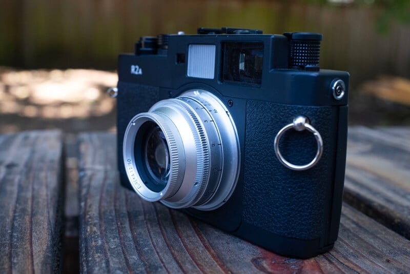 A black and silver rangefinder camera with a prominent lens sits on a weathered wooden surface outdoors, with a blurred background of a fence and greenery.