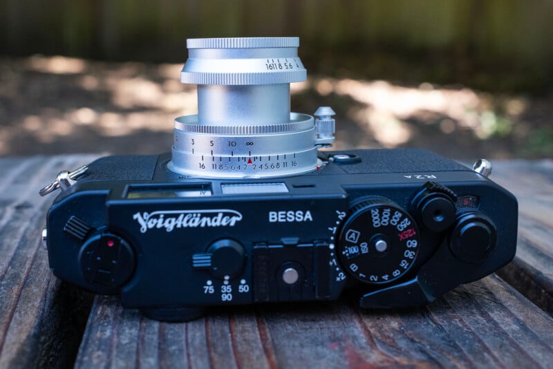 A Voigtländer Bessa R2A film camera with a silver lens is resting on a wooden surface outdoors, with a blurred background of greenery and sunlight.
