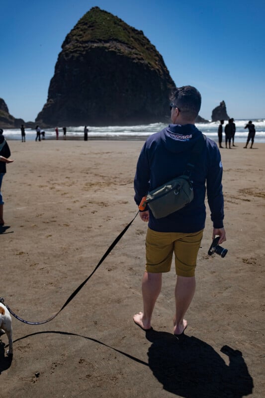 A man stands barefoot on a sandy beach, holding a camera and a dog leash. He faces the ocean and a large rock formation in the distance, under a clear blue sky with other people in the background.
