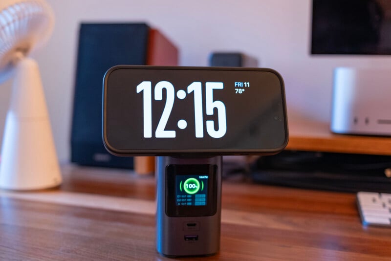 A digital clock displaying 12:15 and 78°F sits on a wooden desk, with a small screen below showing a phone charging at 100%. Electronics and a fan are visible in the background.