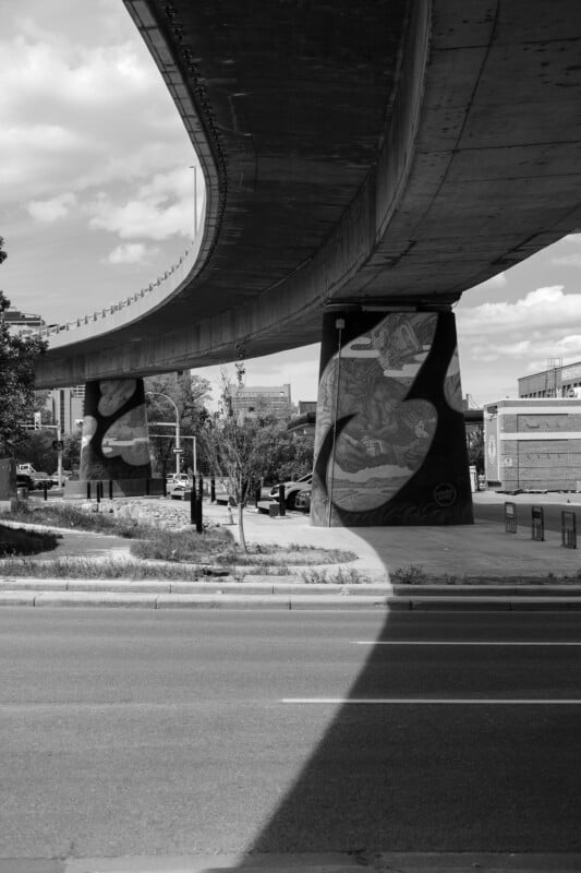 A black and white photo shows a large overpass casting a shadow over a city street. The supporting columns feature murals, and trees and buildings are visible in the background under a partly cloudy sky.