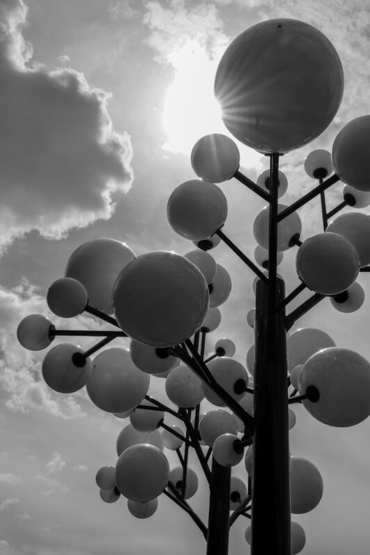 Black and white photo of a modern sculpture with multiple shiny spherical shapes on tall poles, resembling abstract trees, set against a partly cloudy sky with the sun shining through.