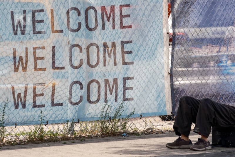 A large banner reading "WELCOME" three times hangs on a chain-link fence. In the foreground, a person sits on the ground with legs outstretched, partly visible from the waist down.