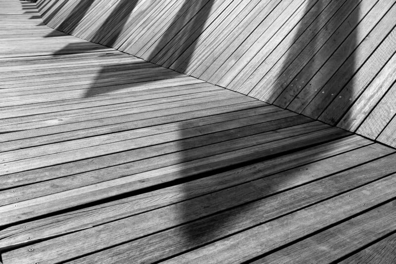 Black and white photo of a wooden boardwalk with diagonal wooden planks rising to form a wall. Shadows of geometric shapes fall across the walkway, creating a pattern on the wood surface.