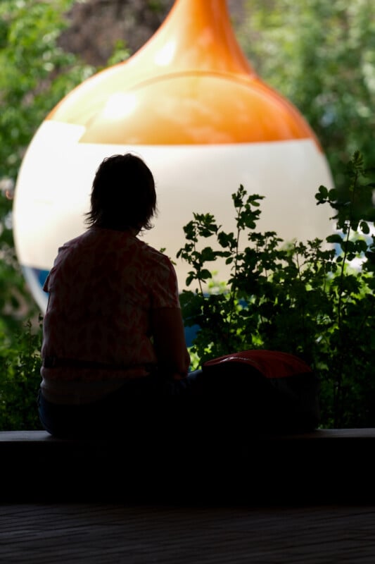 A person sits in shadow on a bench, facing leafy greenery and a large, colorful, blurred object outside, possibly a playground structure or sculpture.