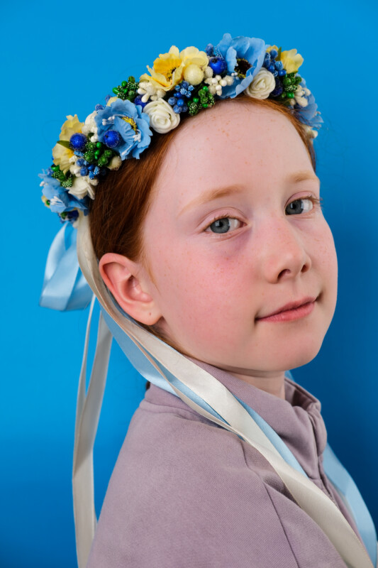 A young girl with red hair wears a colorful floral crown with blue, yellow, and white flowers, and long white and blue ribbons, against a bright blue background. She looks at the camera with a gentle expression.