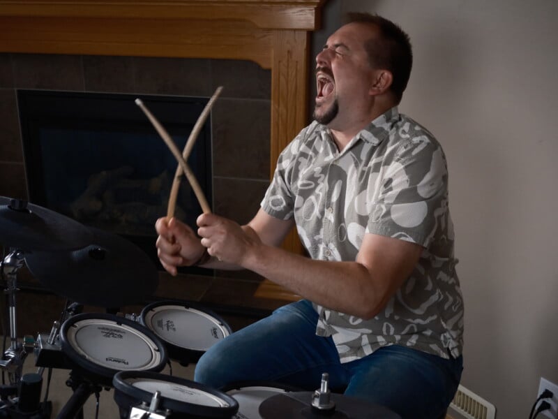 A man wearing a patterned short-sleeve shirt passionately plays an electronic drum set, holding drumsticks crossed in the air and singing or shouting, with a fireplace in the background.