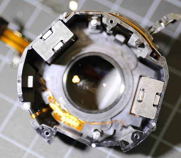 Close-up view of the inner components of a disassembled camera lens, showing the glass lens element, wires, and metallic mechanisms, placed on a grey grid-patterned surface.