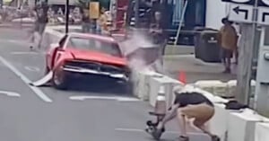 A red car crashes into a concrete barrier on a city street as a photographer crouches nearby, capturing the moment. Dust and debris are visible from the impact, with several bystanders in the background.