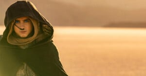 A person with blue eyes and blue markings on their face wears a hooded cloak and scarf, standing in a desert landscape with soft, golden sunlight in the background.
