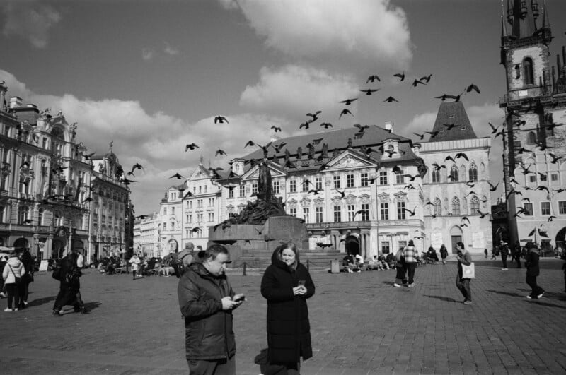 A black-and-white photo of a European square with historic buildings, people walking, and a flock of birds flying in the sky; two people stand in the foreground, bundled in coats.