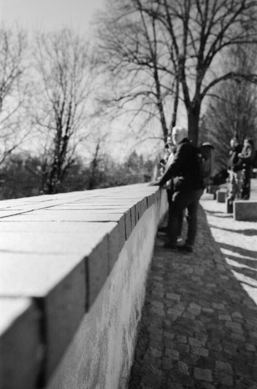 A black and white photo shows people standing by a low stone wall on a cobblestone path, with bare trees in the background under bright sunlight.