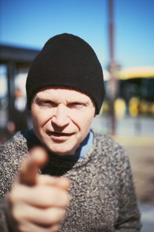 A person wearing a black beanie and a gray sweater points directly at the camera with a serious expression. The background is outdoors with a blurred yellow vehicle and blue sky.