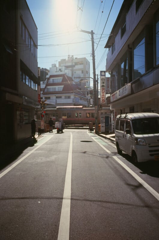 A quiet urban street with sunlight streaming down, a train passing through a railway crossing in the background, and a white van parked on the right side. Buildings line both sides of the street.