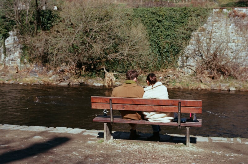 A couple sits closely together on a wooden bench by a river, wrapped warmly in coats and a blanket, facing the water. Leafless trees and a stone wall can be seen in the background.