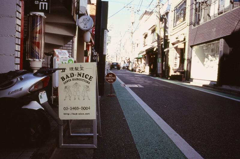 A street scene in Japan shows a sidewalk with a scooter and a barber shop sign reading "BAD-NICE Gents Haircutters" near shops and buildings, with a green bike lane and overhead power lines visible.