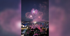Colorful fireworks burst over a harbor at night, illuminating boats on the water and buildings along the shore. The sky is filled with pink and purple lights reflecting on the calm surface below.