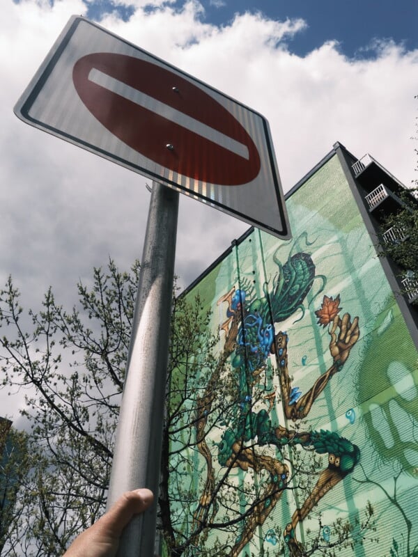 A person holds a no-entry traffic sign in the foreground, with a colorful mural of a robotic figure on a green building wall and trees in the background under a partly cloudy sky.