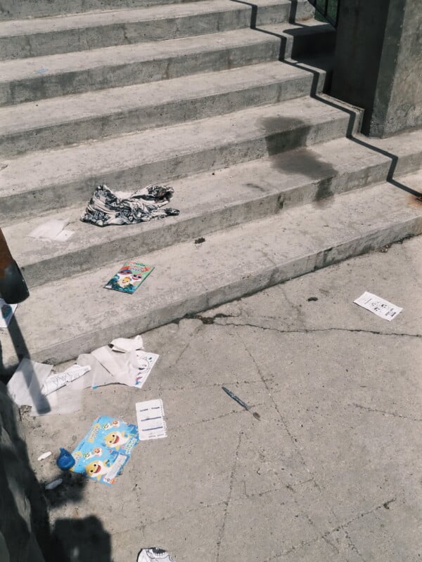 A concrete staircase with scattered litter, including papers, packaging, and clothing, on the steps and the ground below. The scene appears outdoors, in daylight.