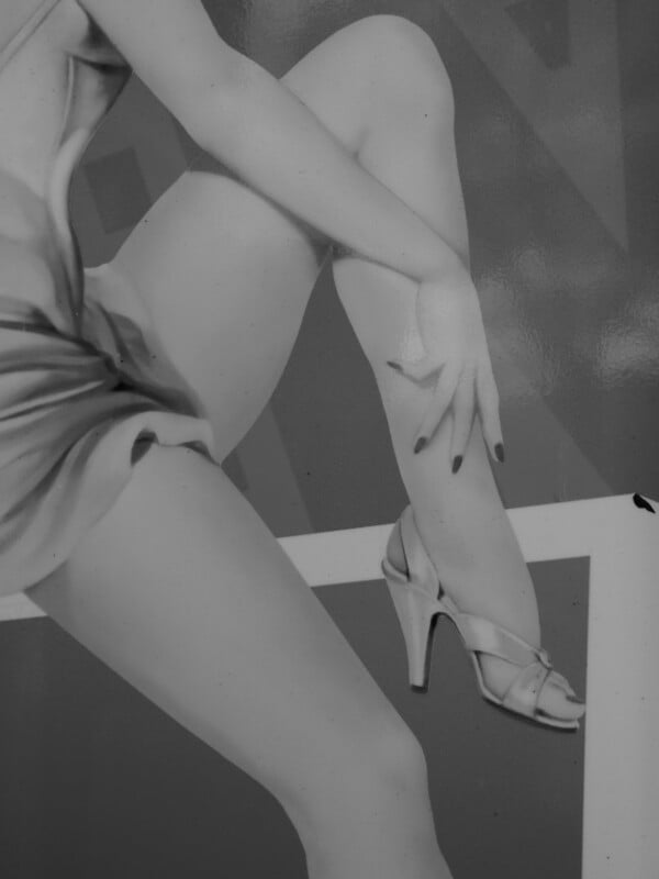 Black and white photo of a person in a short, silky dress sitting with one knee raised, crossing their legs. They wear high-heeled sandals and have one hand resting on their shin, showing manicured nails.