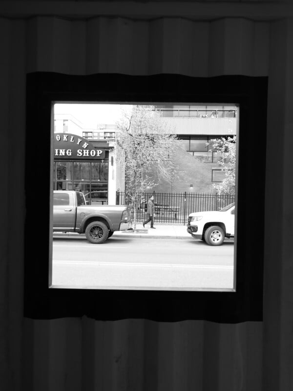 Black and white photo of a city street viewed through a square window. Two parked trucks and a person walking past a building marked "ING SHOP" are visible outside.
