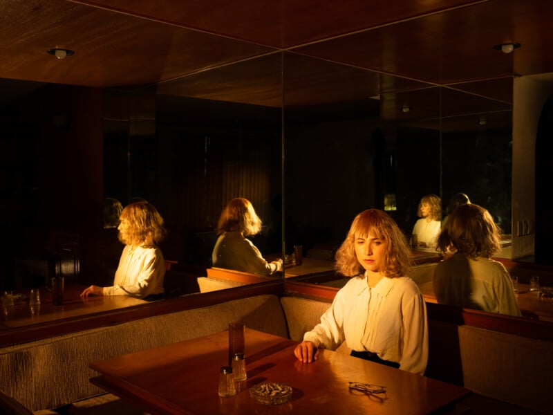 A woman with blonde hair sits alone at a wooden table in a dimly lit room, her reflection appearing multiple times in the mirrors behind her. Warm light emphasizes her contemplative expression. Drinks and an ashtray are on the table.